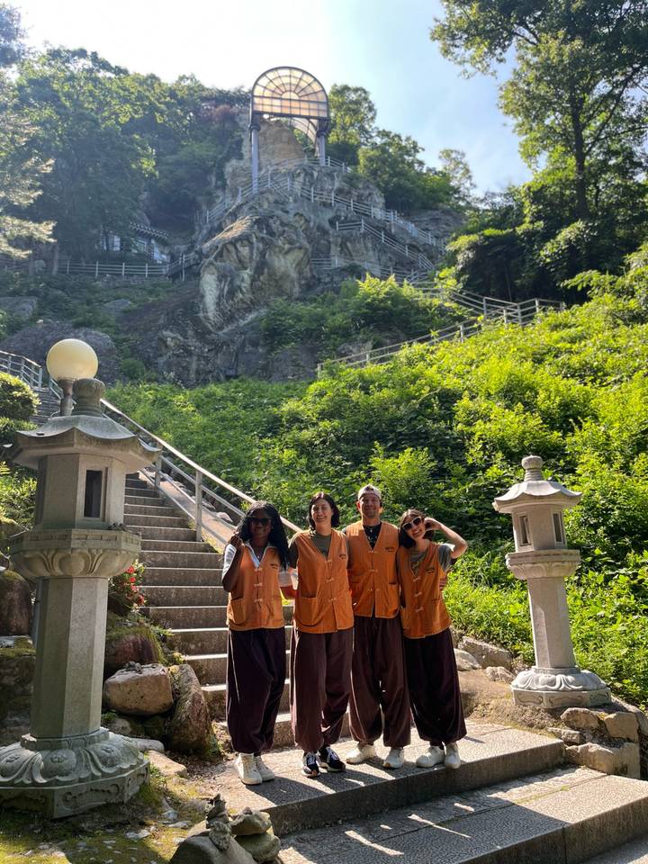 Four volunteers in orange vests stand at the base of a forested temple staircase on a sunny day.