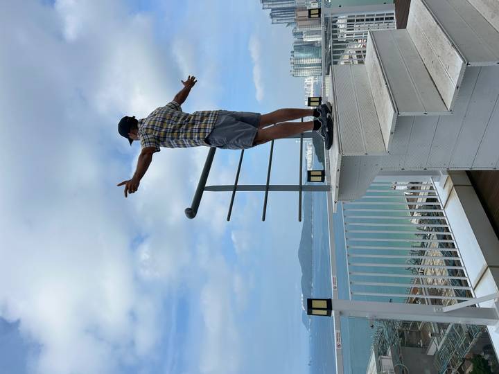 Traveler with outstretched arms enjoys a rooftop coastal view of turquoise waters and city skyline under patchy clouds.