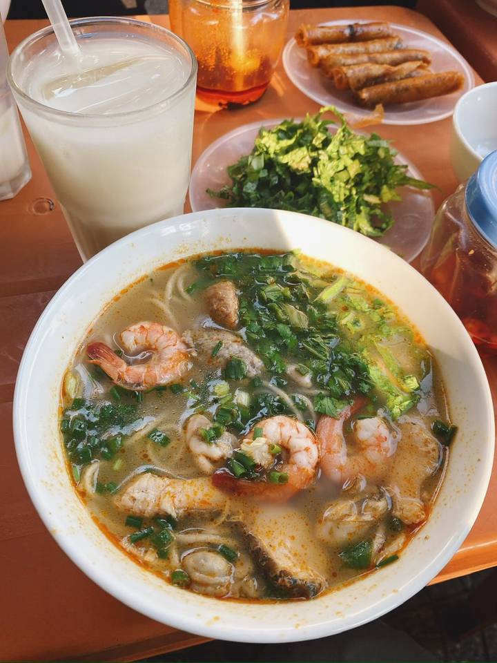 Bowl of aromatic shrimp noodle soup garnished with fresh herbs alongside iced drink on a plastic table.