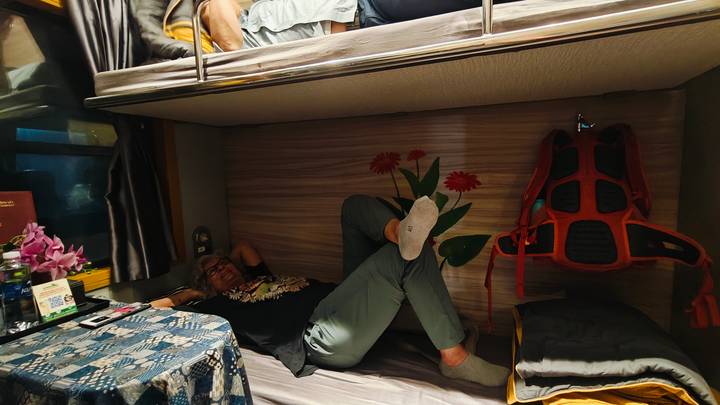 Traveler lounging in lower berth of a sleeper train cabin decorated with floral motif.