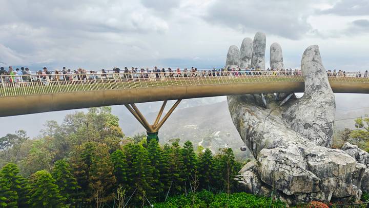 Iconic Golden Bridge held by giant stone hands above lush mountains in Ba Na Hills with crowds walking across.