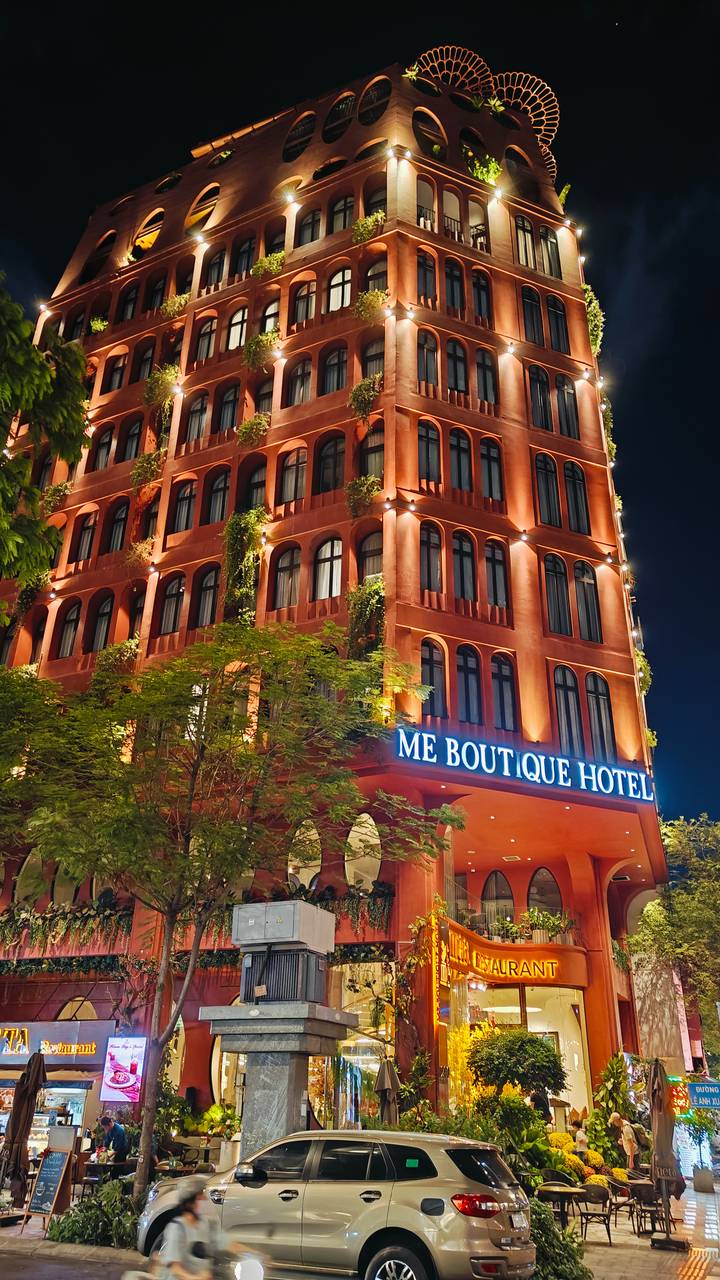 Night view of the illuminated red-orange façade of ME Boutique Hotel framed by leafy trees.