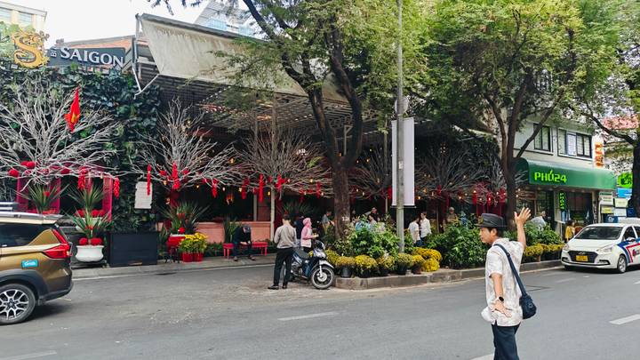 Tree-lined Saigon street with festive red decorations, scooters, and a man hailing a ride.