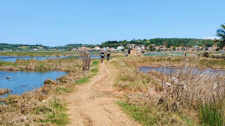Cyclists riding along a narrow dirt path between flooded fields with a rural village beyond.