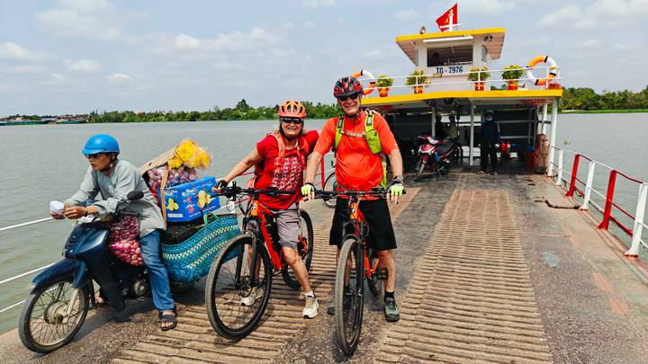 Cyclists with mountain bikes on a small river ferry, another passenger on scooter beside them under bright skies.