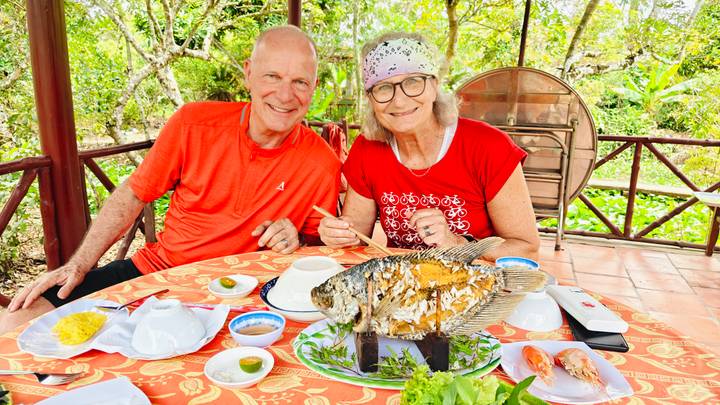 Smiling couple at garden restaurant table with whole grilled fish and Vietnamese dishes laid out.