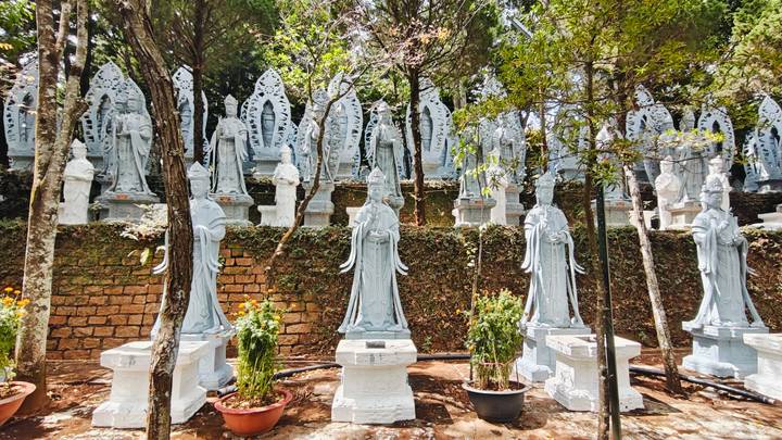 Terraced rows of white stone Quan Âm statues set against a brick retaining wall and dappled forest light.