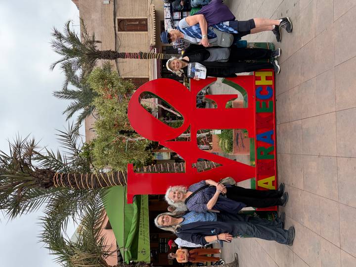 Tourists pose beside a large red LOVE Marrakesh sign surrounded by palm trees.