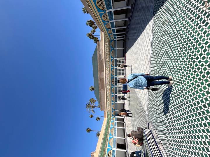 Visitors walk across the patterned courtyard of Bahia Palace in Marrakesh under a deep blue sky.