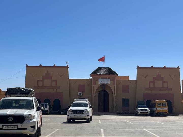 An adobe-style Moroccan public building with a flag and parked vehicles under a clear sky.