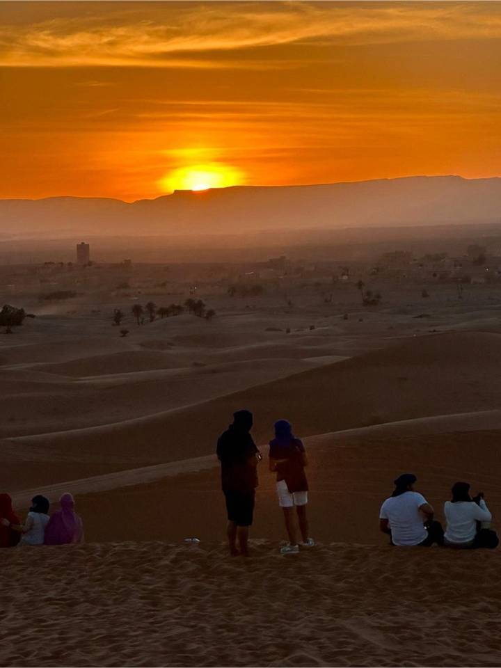 Sahara sunset over distant town with two robed figures on dune foreground.
