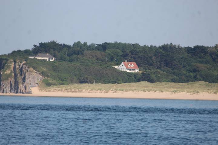 Coastal scene of sandy beach and distant white house nestled in wooded headland across calm blue water.