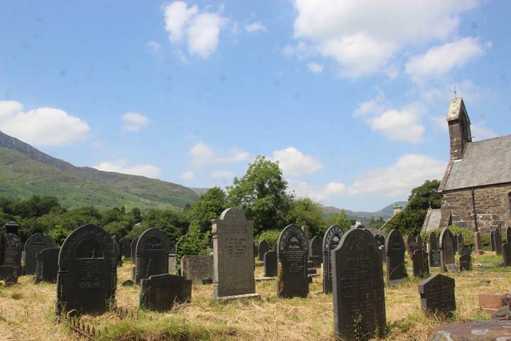 Old hillside cemetery with stone headstones beneath partly cloudy blue sky and rolling green mountains.