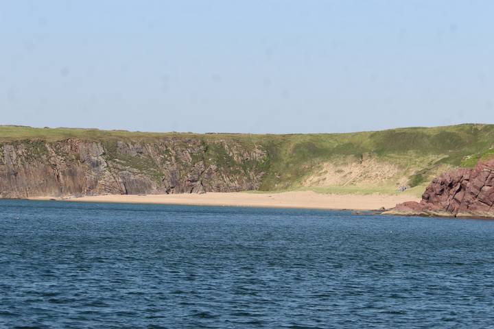 Remote sandy cove bordered by rugged green cliffs beside open blue sea.