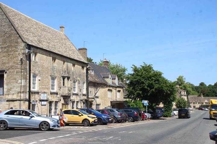 Typical Cotswolds village street lined with honey-stone buildings and parked cars on a sunny day.