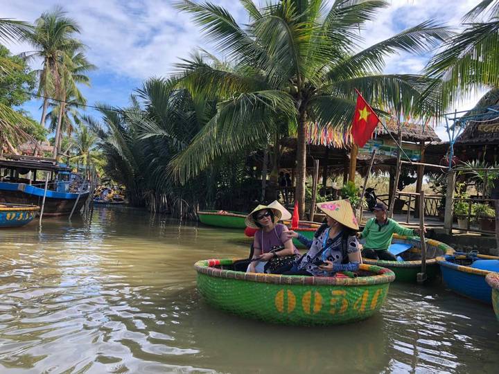 Tourists wearing conical hats ride colorful round basket boats along a palm-lined canal with Vietnamese flag flying
