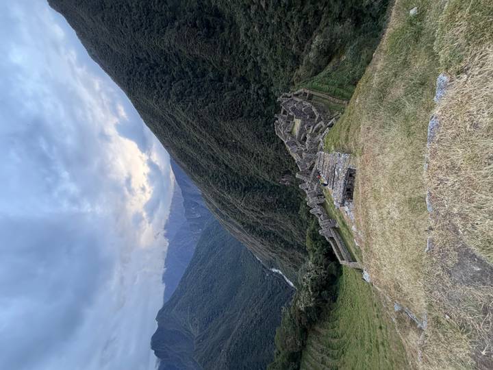 Stone Inca ruins cascade down a steep grassy ridge above a remote cloud-filled valley at dusk