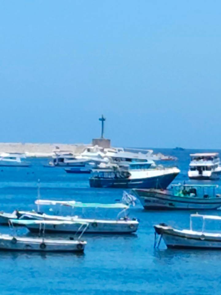 Blurry view of small boats and yachts on bright blue water near a low harbour wall and a tiny lighthouse