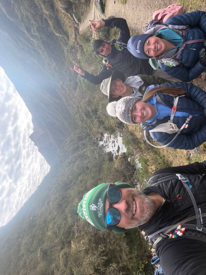 Smiling hikers beside a mountain stream with layered clouds and high Andean peaks in the distance