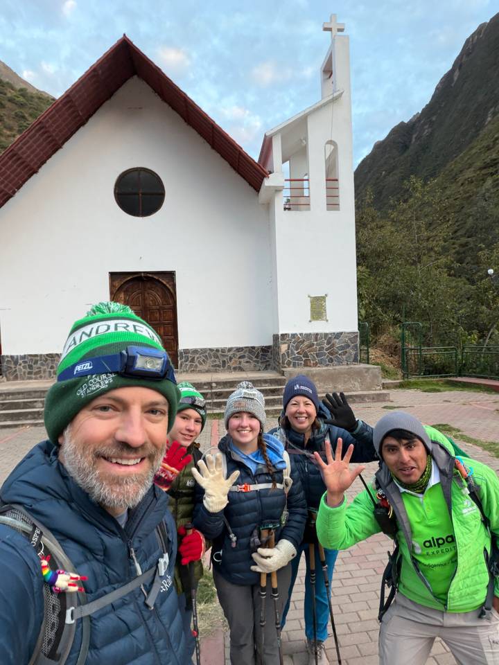 Selfie of trekkers waving outside a small white chapel in a mountain village