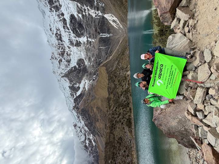 A trekking group displays an Alpaca Expeditions banner beside a turquoise lake beneath snowy Andean peaks.