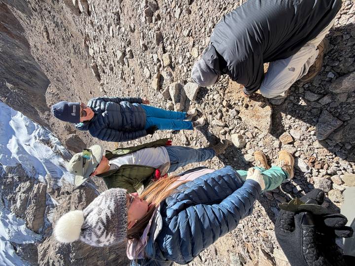 Trekkers gather on a rocky slope while a guide demonstrates geology near a glacier.