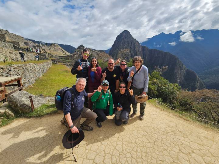 A smiling tour group poses at the classic Machu Picchu viewpoint with Huayna Picchu towering behind.