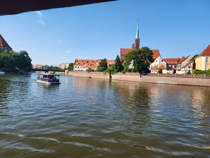 Tour boat cruises a calm river past red-roofed churches and leafy embankments on a bright day.