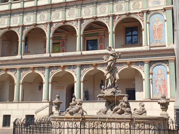 Renaissance town hall facade with colorful frescoed arches and an ornate fountain in front under bright sunlight.