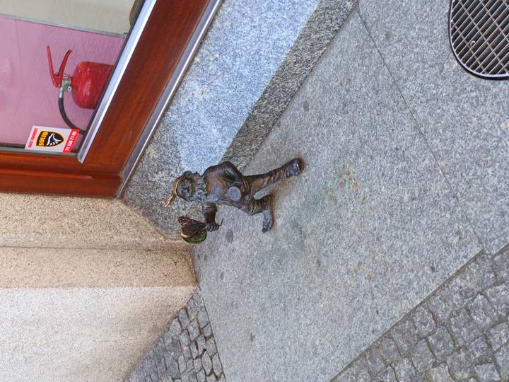 Bronze gnome statue holding a hat and coin on a granite sidewalk beside a shop window.