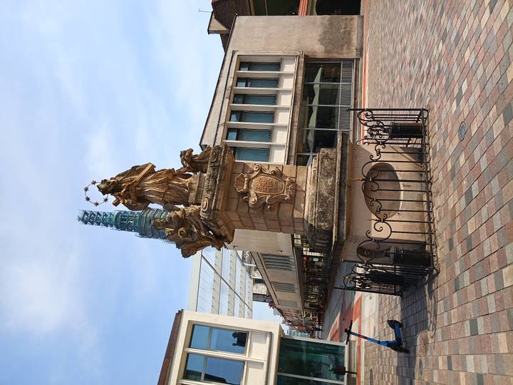 Baroque column monument topped by a crowned saint surrounded by stars on a paved plaza beside modern buildings.