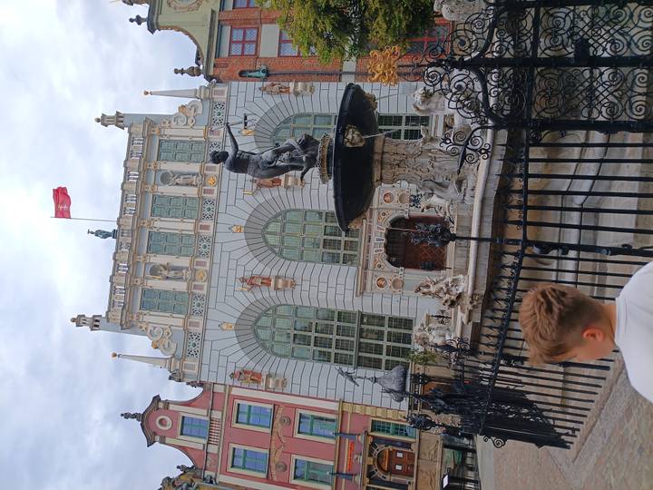 Neptune Fountain in front of the ornate Artus Court building flying the Polish flag, with a child observing in the foreground.