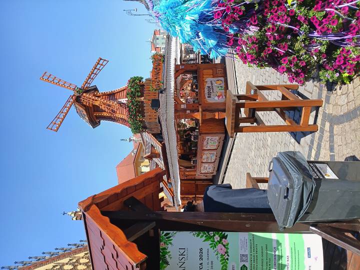 Wooden windmill-themed market stall on a sunny square, with tables, flowers and a trash bin in the foreground.
