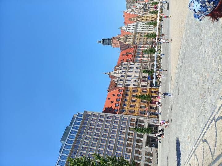Wide view of colorful historic townhouses bordering a spacious cobbled market square filled with strolling visitors under a clear blue sky.