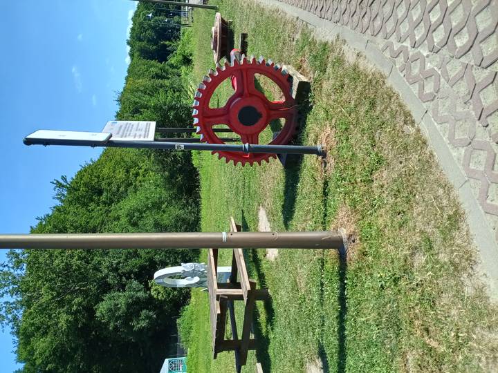 Outdoor display featuring a large red gear wheel and machinery parts beside a grassy path and wooded backdrop on a sunny day.