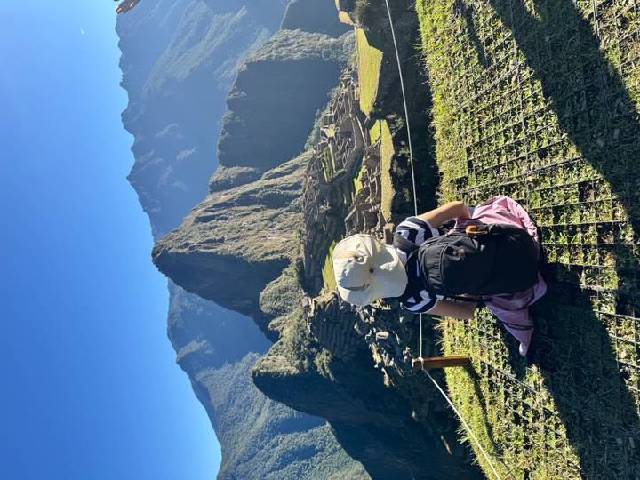 Traveler sitting on a grassy ledge overlooking the ancient stone terraces of Machu Picchu with dramatic peaks behind.
