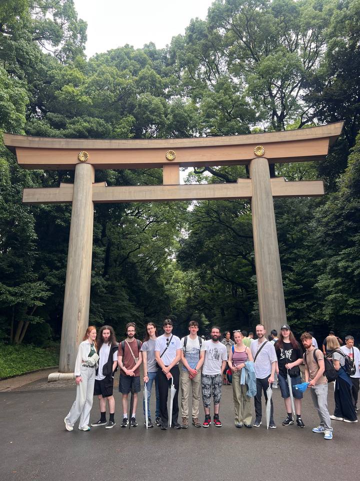 Large travel group stands beneath a massive wooden torii gate in a lush forest setting.
