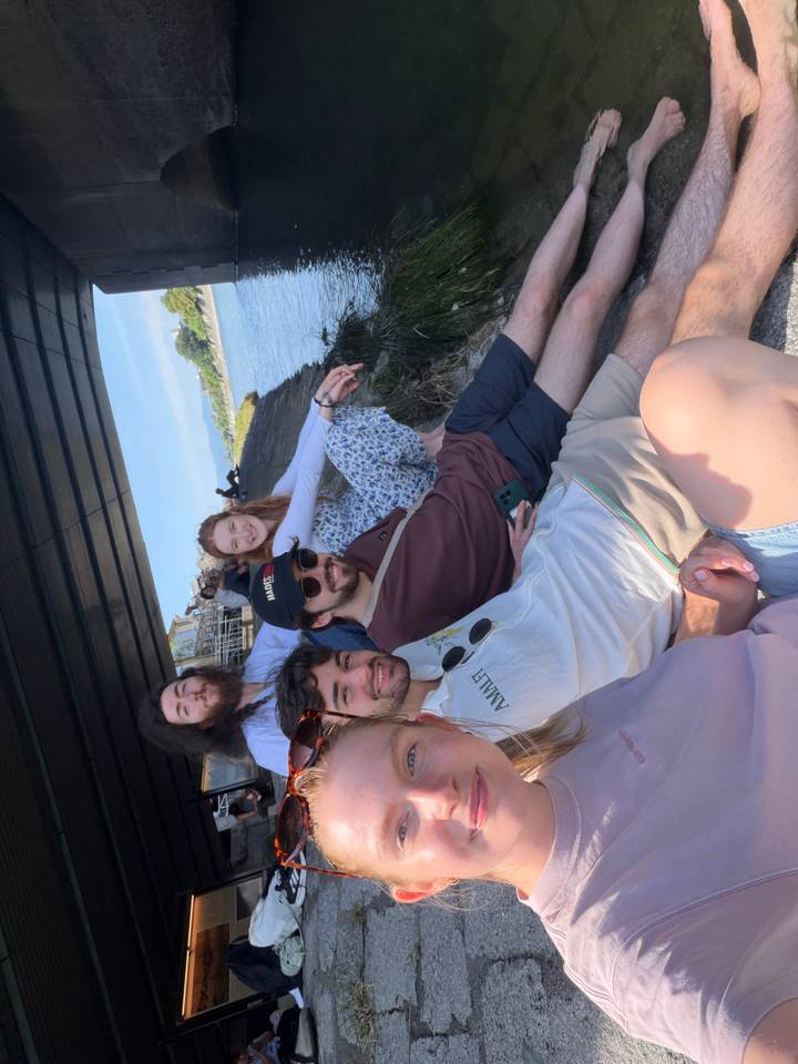 Group relaxes on a riverbank under a bridge enjoying the afternoon sun with cityscape behind.
