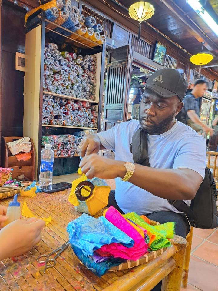 Traveller crafts a colourful mask with paper and glue inside a bustling market workshop.