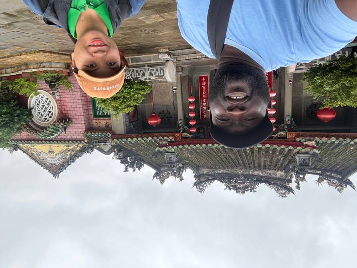 Two travelers pose in front of an ornate temple with green tiled roof and red lanterns on a cloudy day.
