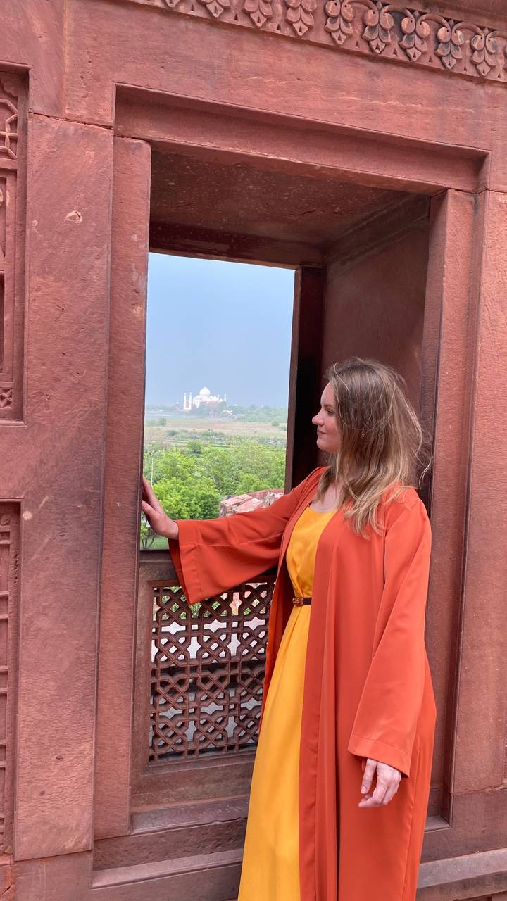 A traveler in an orange jacket gazes through a red-sandstone window toward the distant Taj Mahal across green plains.