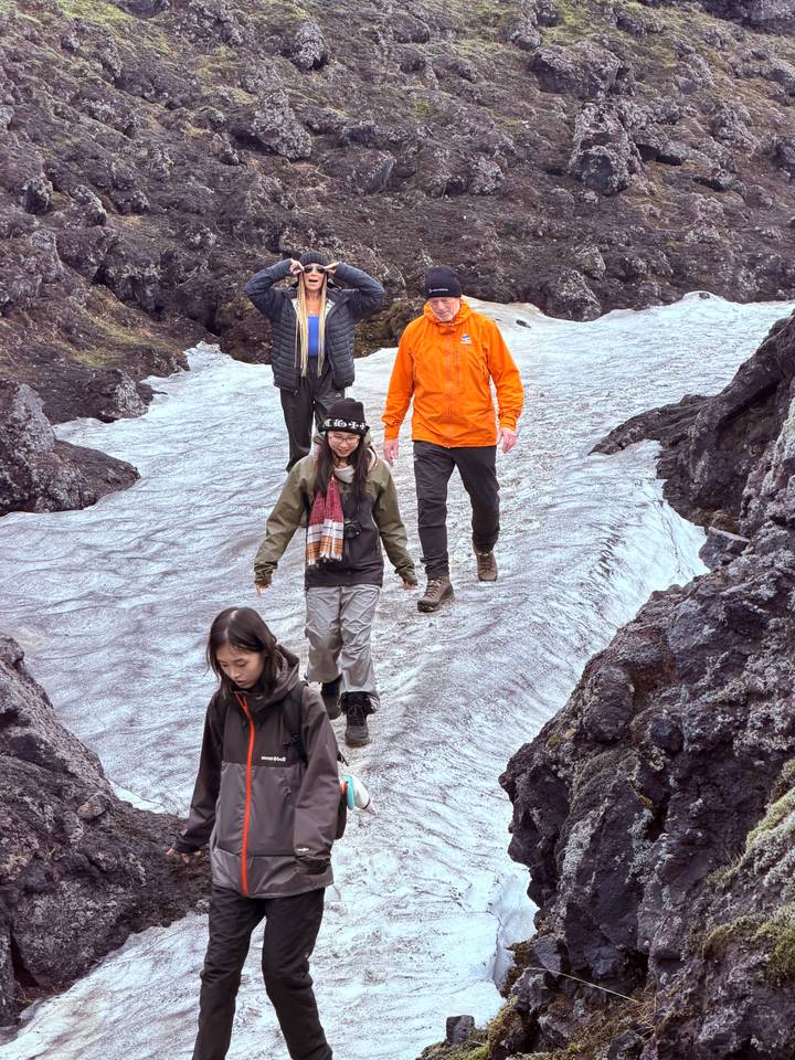Four hikers carefully making their way across a narrow strip of ice and snow between dark volcanic rocks