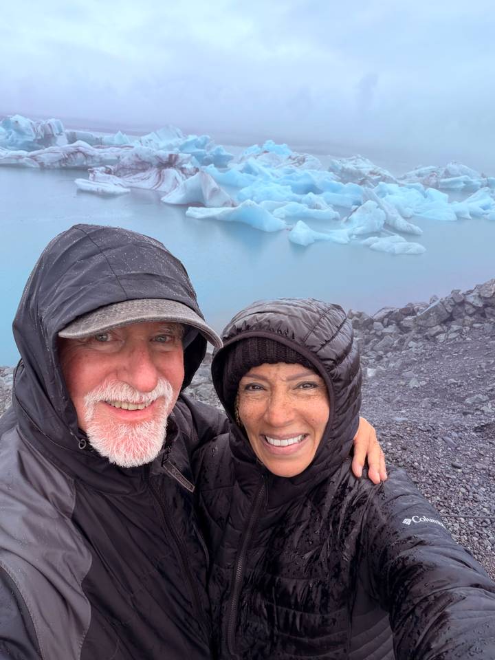 Couple in rain jackets smiling beside a blue glacial lagoon dotted with icebergs