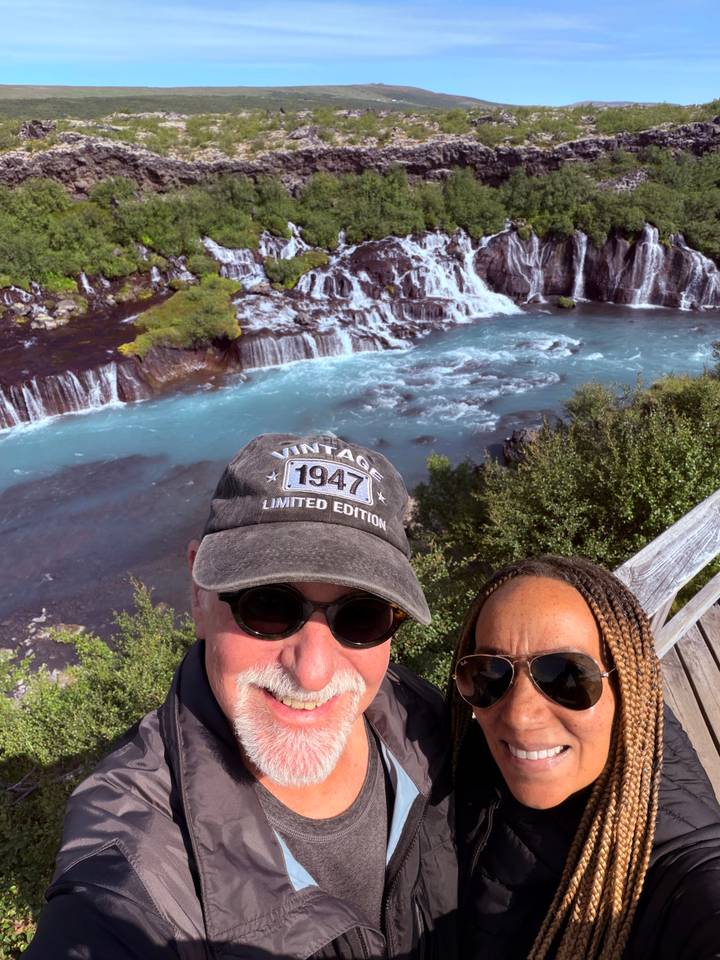 Selfie overlooking a wide blue cascade flowing through dark lava fields and shrubs