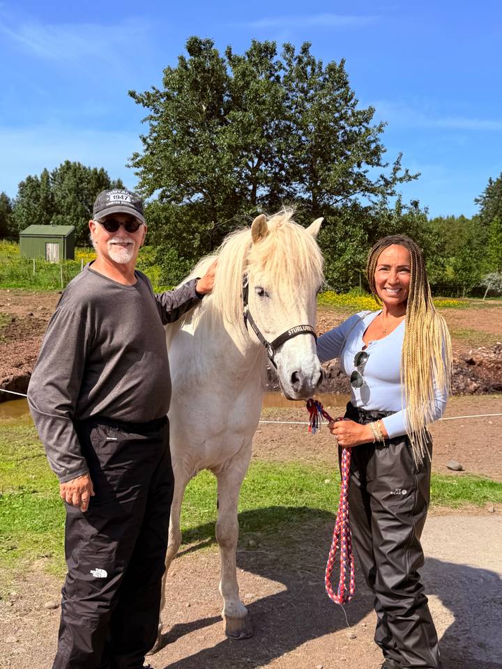 Man and woman posing with a friendly white Icelandic horse in a sunlit pasture