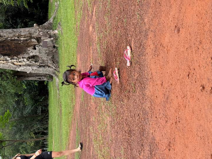 Small girl in bright clothes playing with dirt on a red clay ground in a forest clearing
