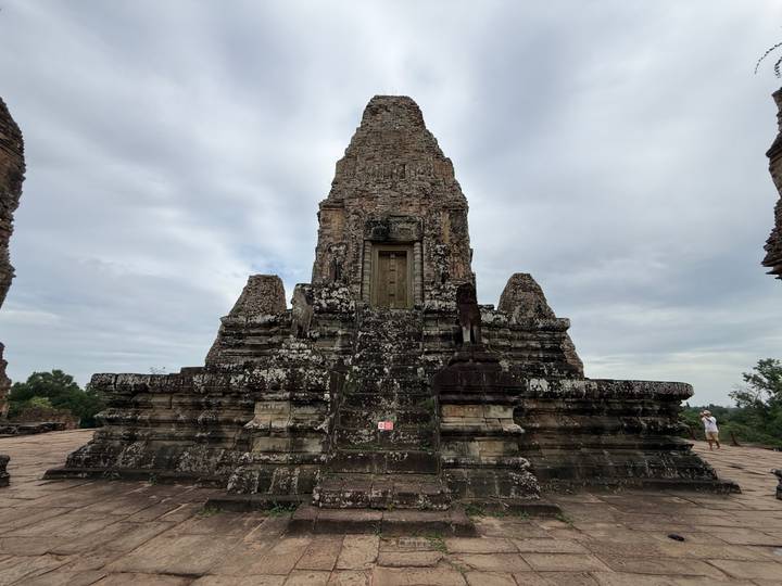 Tall stone temple tower on a platform against a cloudy sky, visitor seen in the distance