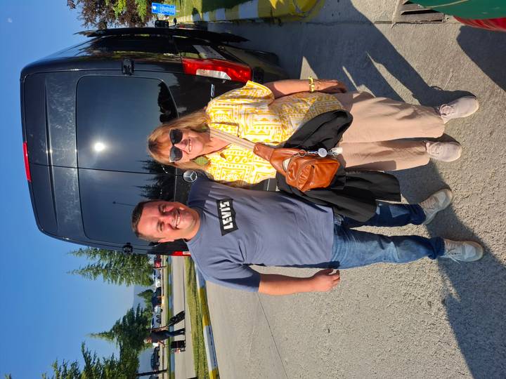 A smiling tourist and driver pose together in front of a black passenger van on a sunny morning.