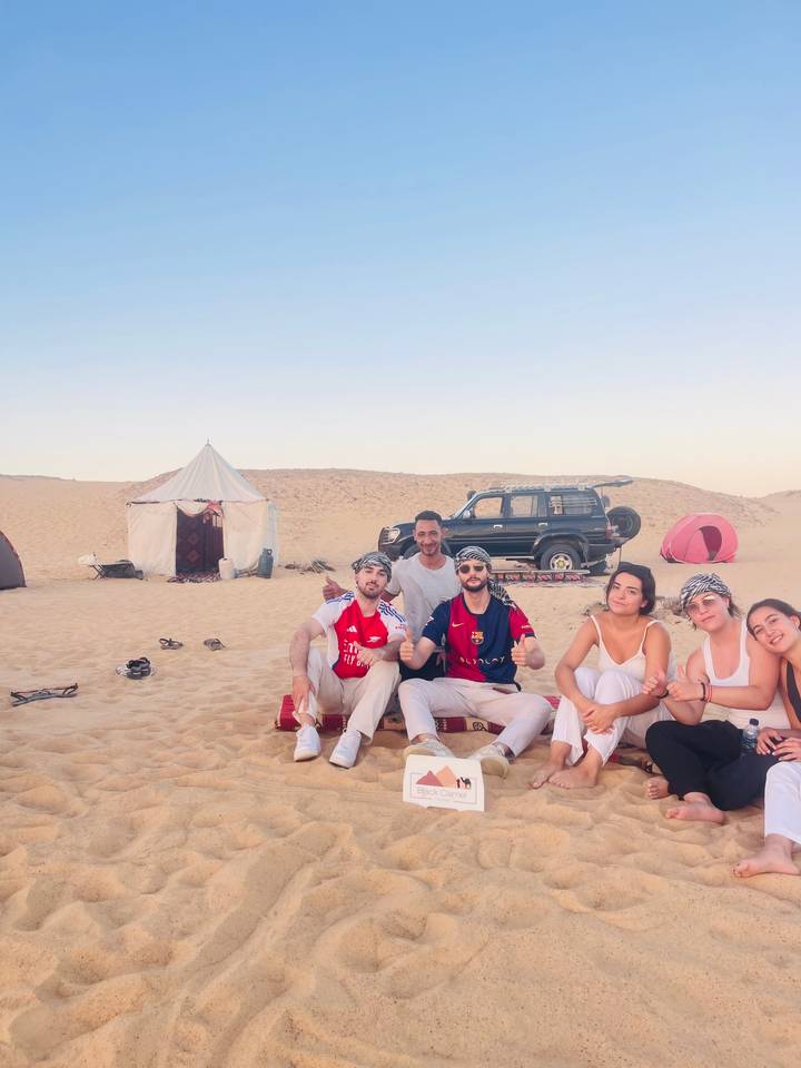 Group of friends relax on desert sand near tents and a 4x4 jeep during a Sahara camp.