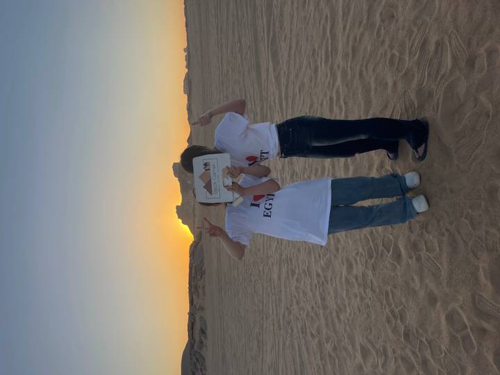 Two young women pose at sunset in the desert holding a tour sign that hides their faces.